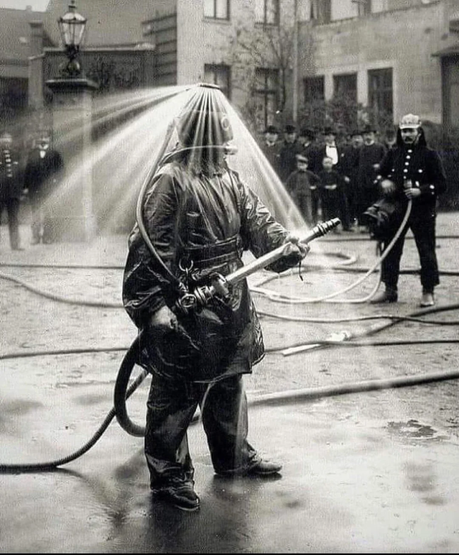 A firefighter in an old-fashioned protective suit is spraying water from a hose. Jets of water shoot out from the helmet in all directions. People in uniforms watch in the background. The photo is black and white.