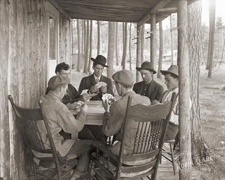 Six men in hats sit around a table playing cards on a wooden porch surrounded by trees. Two men are in rocking chairs, and the atmosphere appears relaxed and rustic.