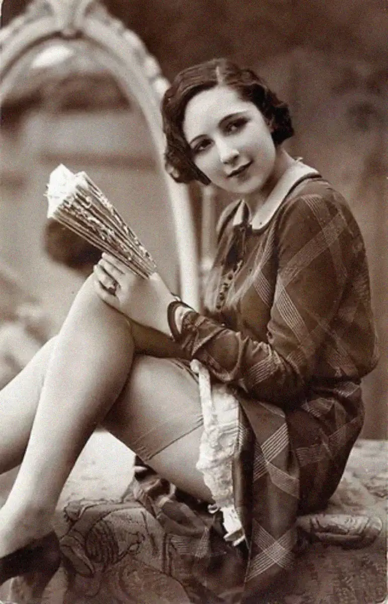 A woman in vintage attire sits with one leg crossed, holding a fan and smiling at the camera. She has short wavy hair and wears a plaid dress, with a decorative mirror in the background. The image is sepia-toned.