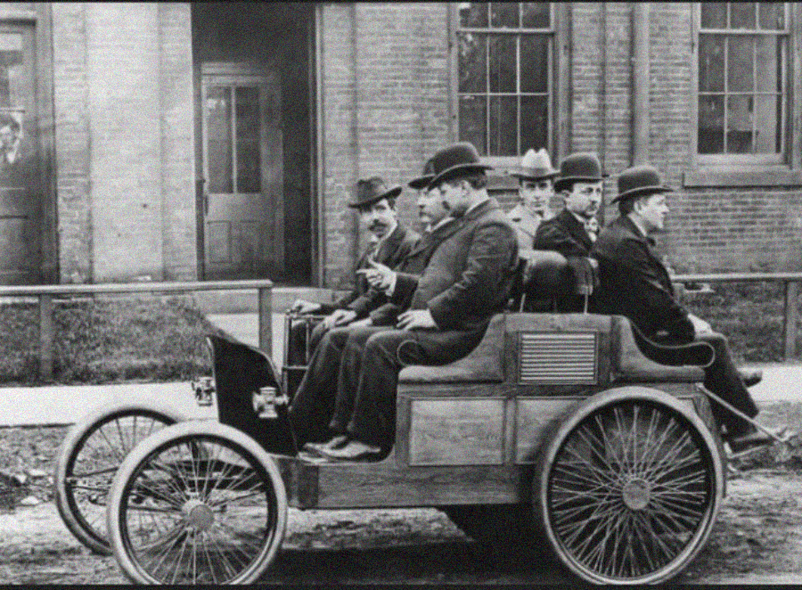 A group of men and one woman, all wearing early 1900s attire and hats, sit in an old-fashioned open automobile in front of a brick building.