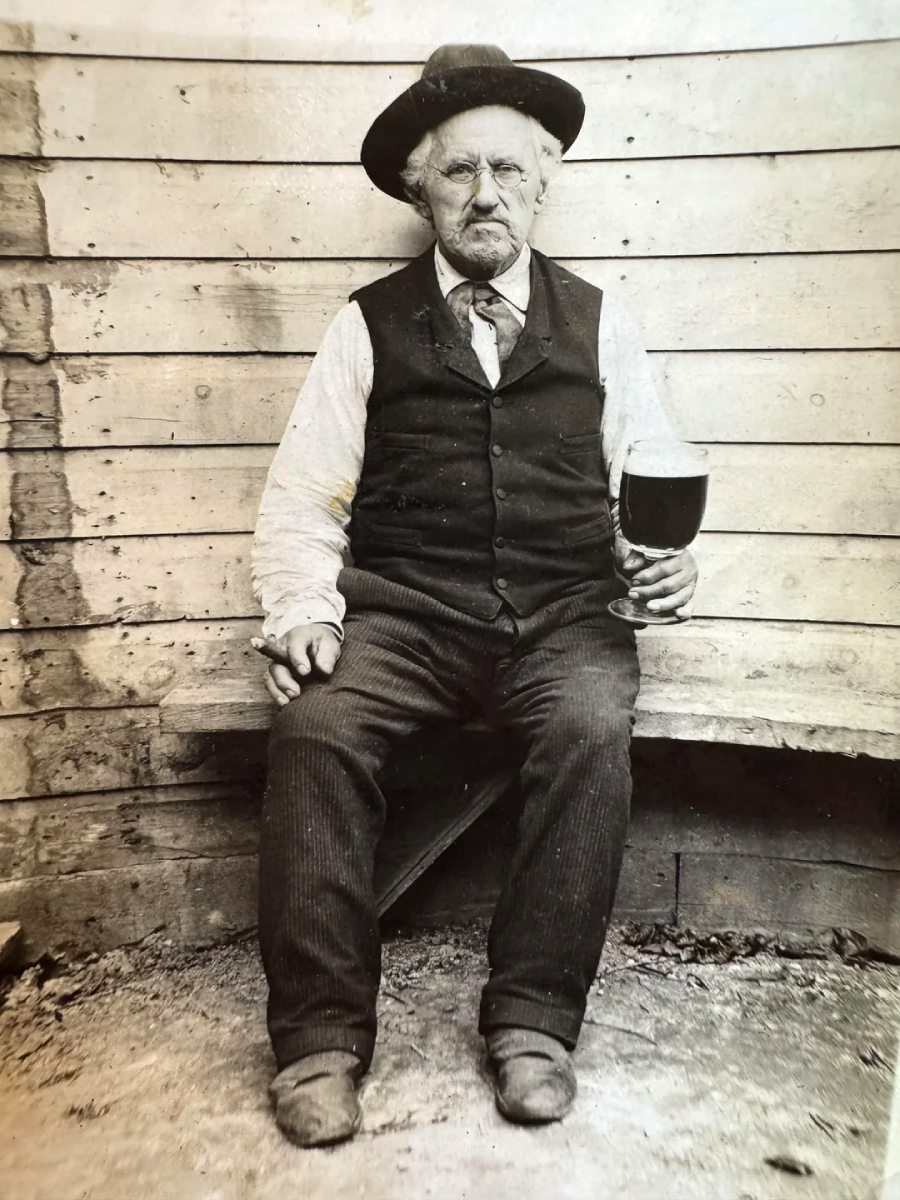 An elderly man with a mustache, wearing a vest, tie, and hat, sits on a wooden bench against a plank wall, holding a large glass of dark liquid and looking at the camera with a serious expression.