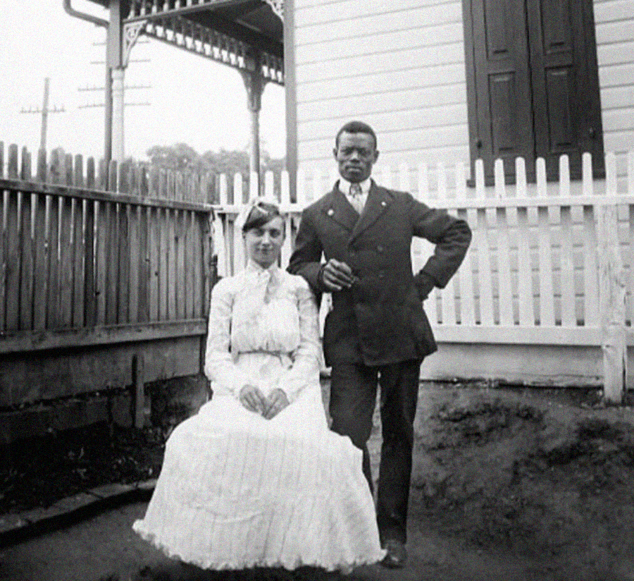 A Black man in a suit stands beside a seated woman in a white dress, both posing for a photo in front of a wooden fence and house with Victorian-style trim.