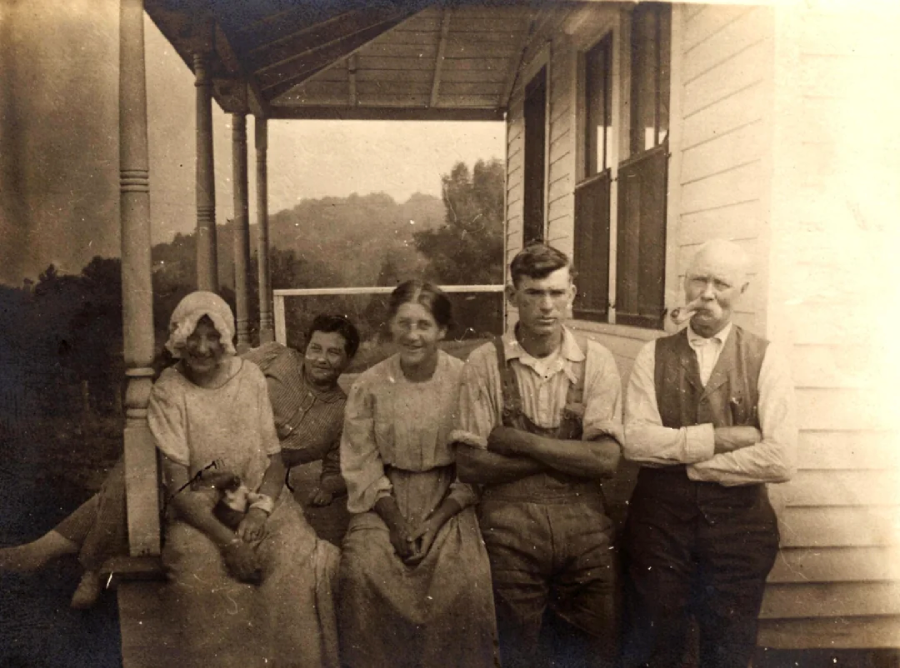 Five people sit and stand on a porch in front of a wooden house. Three women in dresses sit on the porch railing, while a young man and an older man stand beside them. The scene appears old-fashioned and sepia-toned.