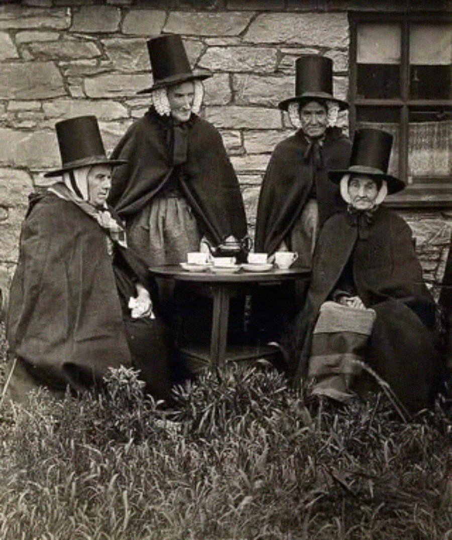 Four women wearing traditional Welsh costumes with tall hats and capes sit and stand around a small round table set for tea outside a stone building with a window. Grass and plants are visible in the foreground.