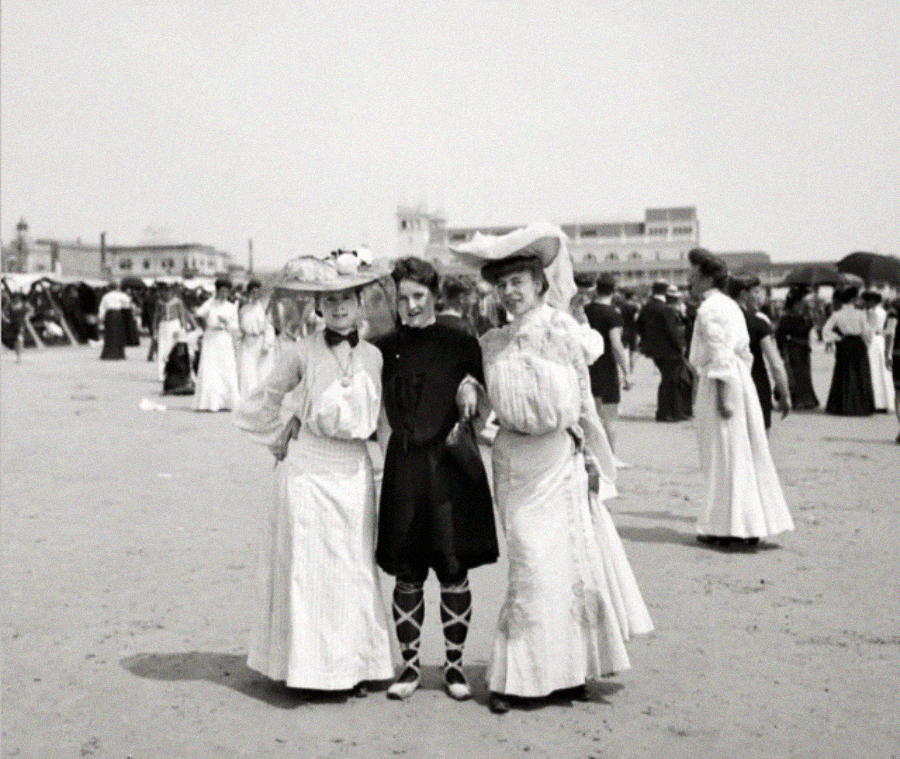Three women stand on a beach, two wearing long skirts, blouses, and wide-brimmed hats, and one in a darker outfit with lace-up boots. Other people in similar attire are visible in the background. The scene appears historical.