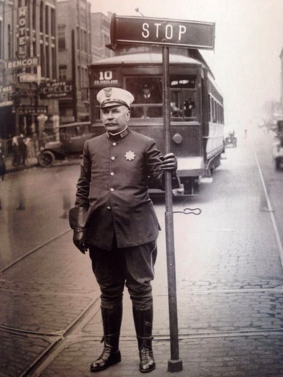 A vintage black-and-white photo shows a uniformed traffic officer holding a tall sign that reads “STOP” on a city street. A streetcar and several buildings are visible in the background.