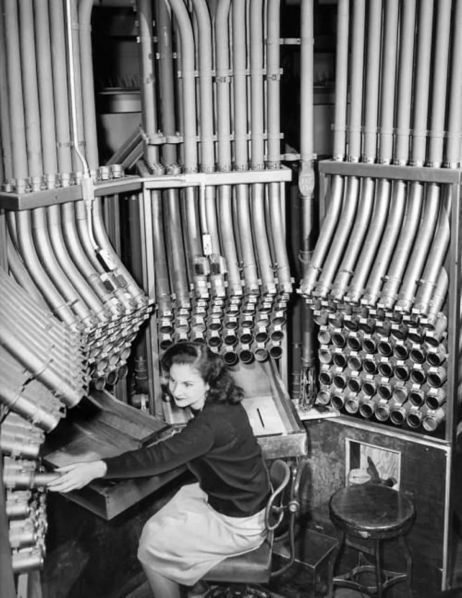 A woman sits at a desk surrounded by numerous rows of metal tubes and pipes, likely part of an old communication or switchboard system. She is looking toward the camera while working.