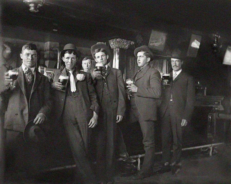 Six men in vintage suits and hats stand in a bar holding up glasses of dark beer, posing for the camera. Framed pictures and a mirror hang on the wall behind them in the dimly lit room.