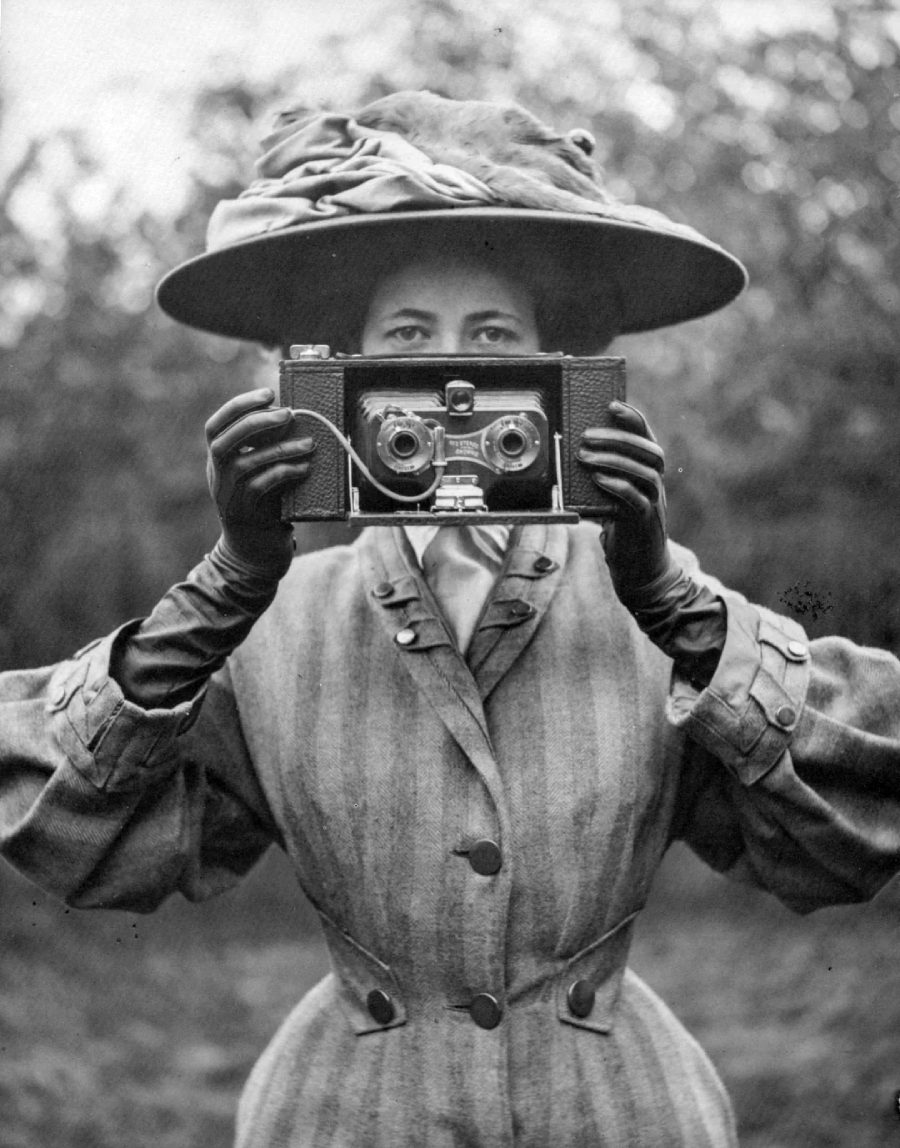 A woman in vintage clothing and a large hat holds an old-fashioned camera up to her face, partially obscuring it. The background is blurred foliage. The image appears to be black and white.