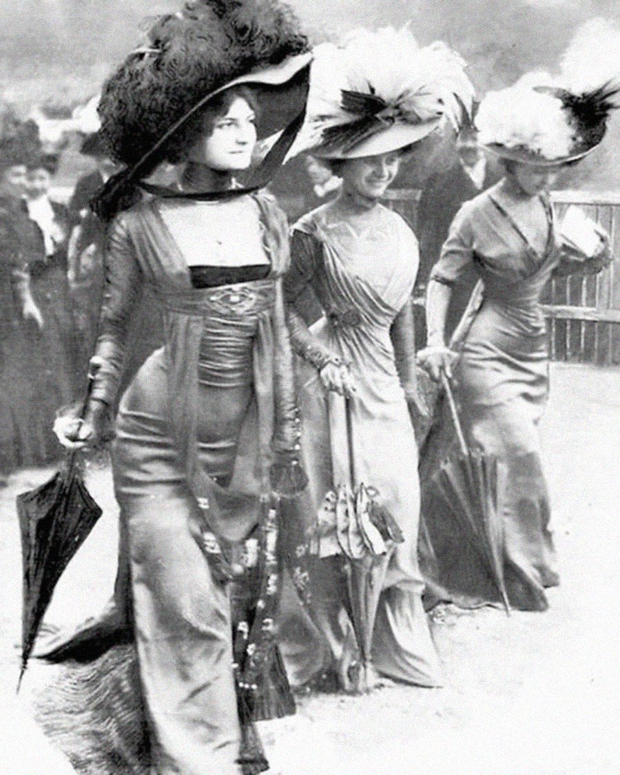 Three women in elaborate Edwardian dresses and large feathered hats walk together, each holding a parasol. The photo is black and white, with other people and a fenced area in the blurred background.
