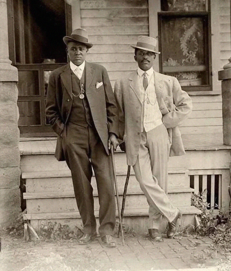 Two stylishly dressed Black men from the early 1900s stand on a porch. Both wear suits, vests, ties, hats, and pocket watches; one leans on a cane while the other stands with his hand in his pocket.