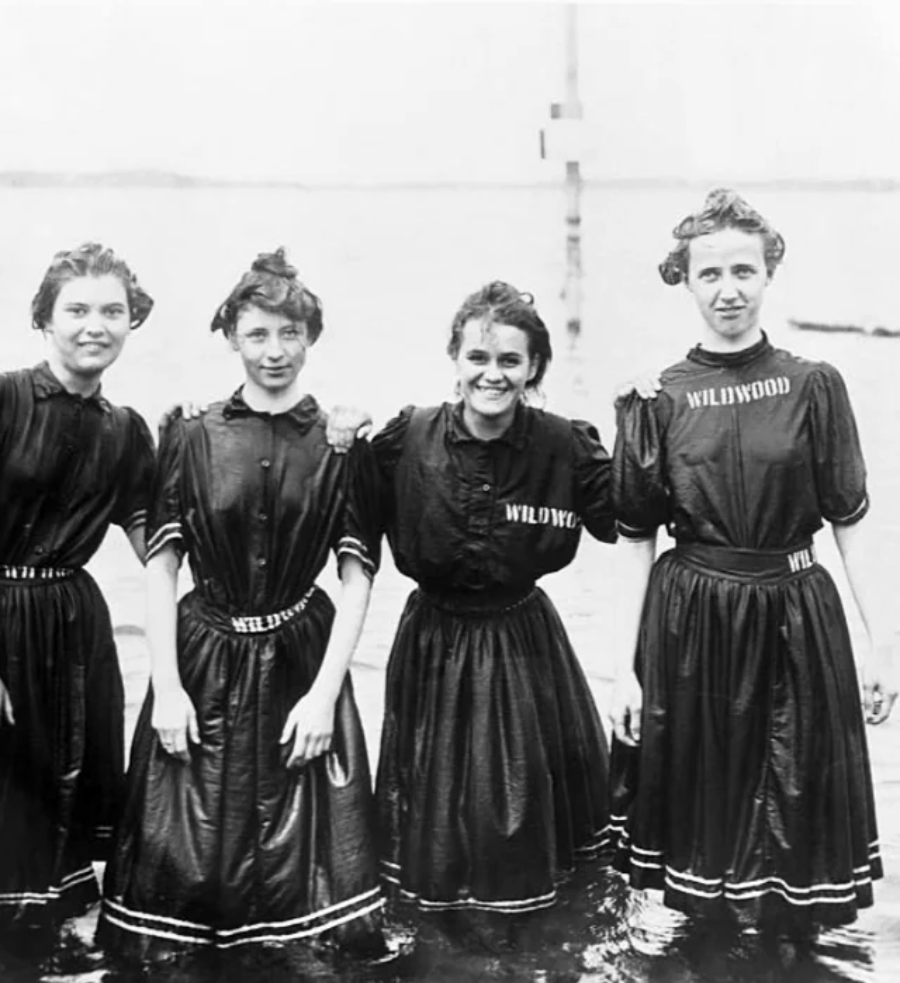 Four women in matching dark, old-fashioned swimsuits labeled "Wildwood" stand together in shallow water, smiling at the camera, with a blurred shoreline in the background.