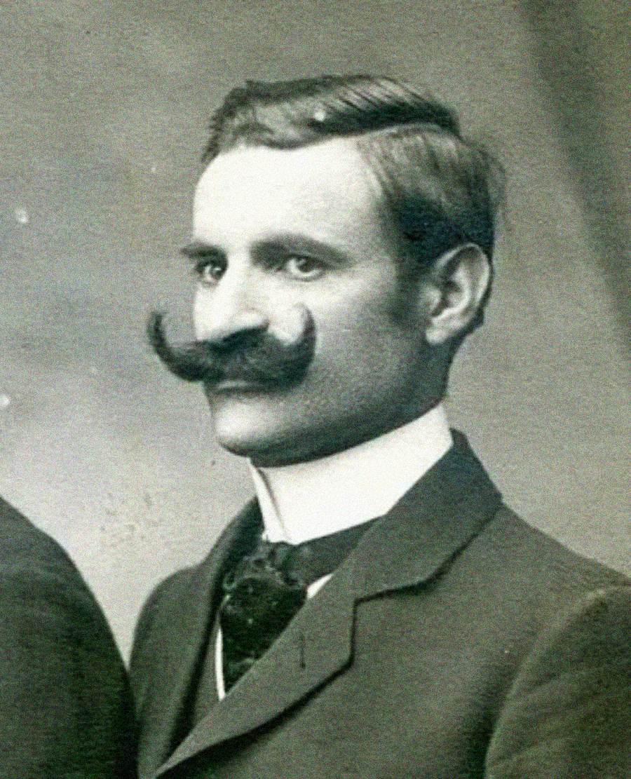 A vintage black-and-white portrait of a man with neatly parted hair, a large, curled mustache, and wearing a suit with a high-collared shirt and tie.