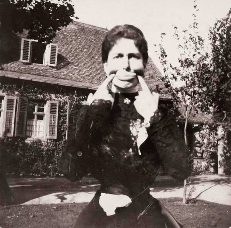 A woman in Victorian-era clothing stands outside in front of a house, pulling the corners of her mouth into a wide smile with her fingers for the camera. Trees and bushes are visible in the background.