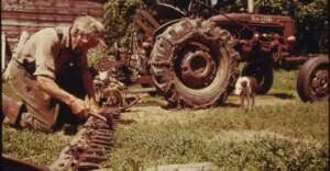 An elderly man kneels on grass, repairing a long metal tool beside a vintage tractor. A small dog stands nearby, and a wooden shed is in the background. The scene is outdoors on a sunny day.