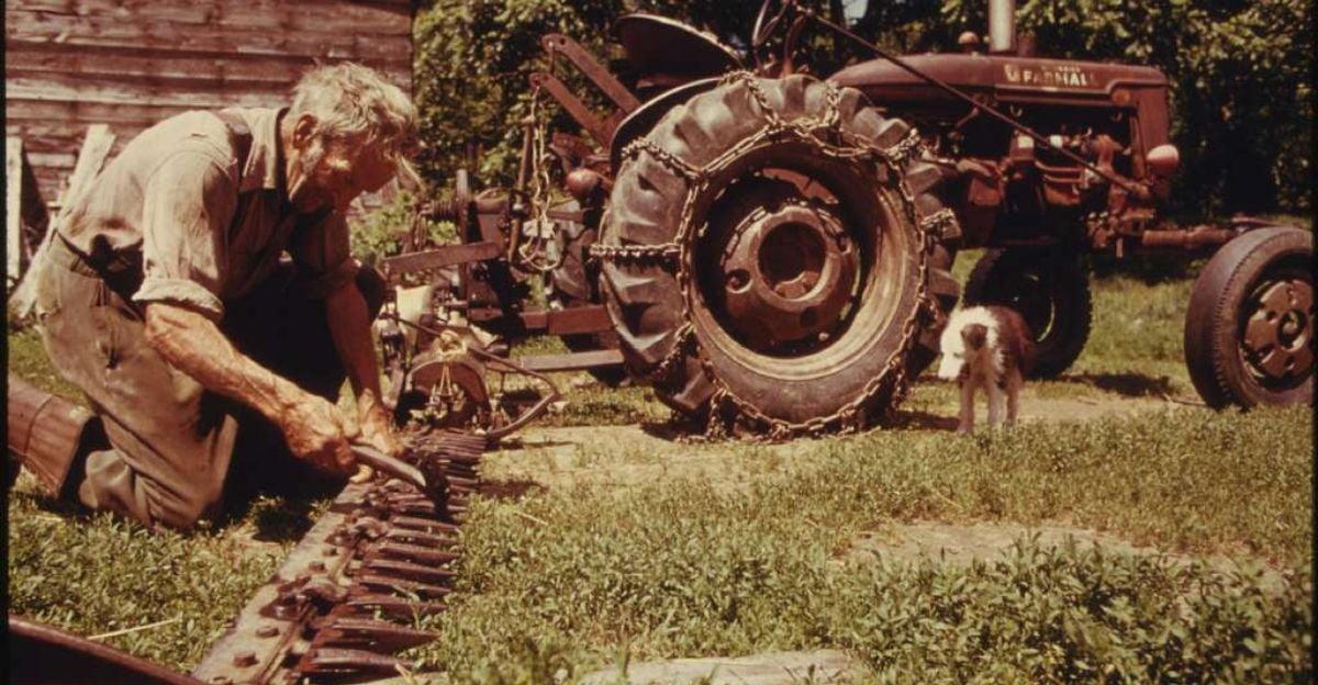 An elderly man kneels on grass, repairing a long metal tool beside a vintage tractor. A small dog stands nearby, and a wooden shed is in the background. The scene is outdoors on a sunny day.
