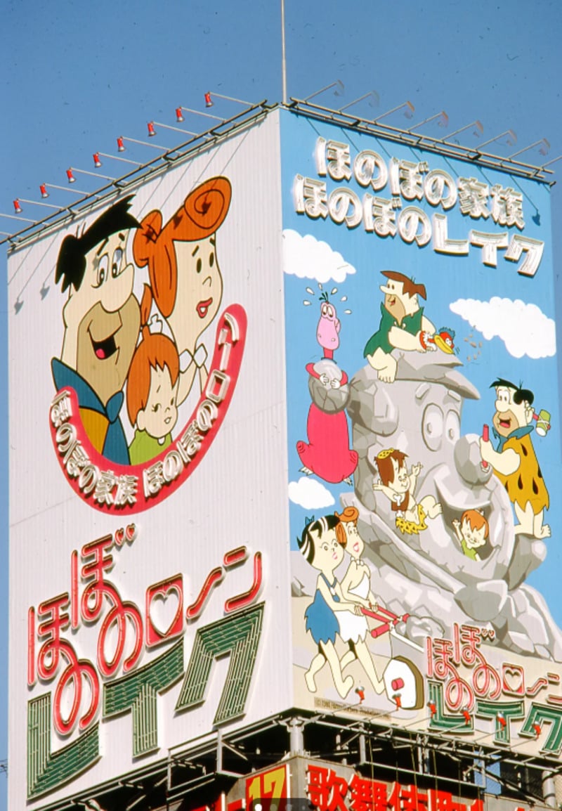 A large Japanese billboard featuring cartoon characters from The Flintstones, including Fred, Wilma, Pebbles, and Dino, with Japanese text and colorful illustrations.