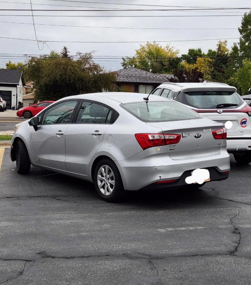A silver sedan is parked awkwardly, occupying two spaces in a parking lot. A white SUV is parked nearby. Trees, houses, and power lines are visible in the background. The license plate is blurred.