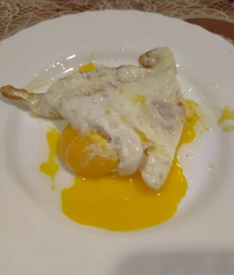 A fried egg with a runny yolk is served on a white plate, with some yolk spilled onto the plate. The egg white is slightly folded and unevenly cooked.