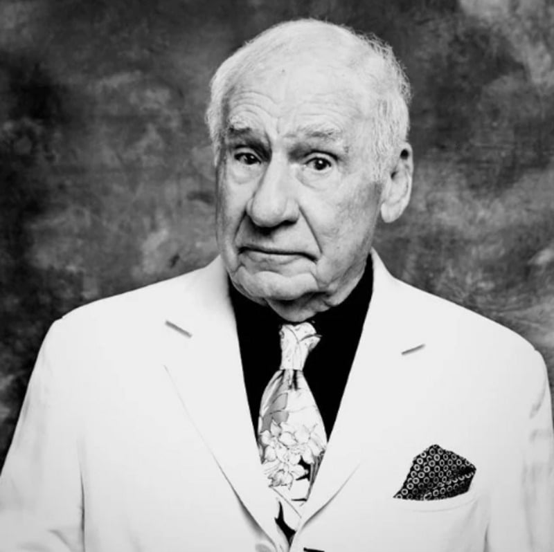An older man with white hair wears a light-colored suit, patterned tie, and pocket square. He stands against a textured backdrop, looking directly at the camera with a slightly raised eyebrow. The photo is black and white.