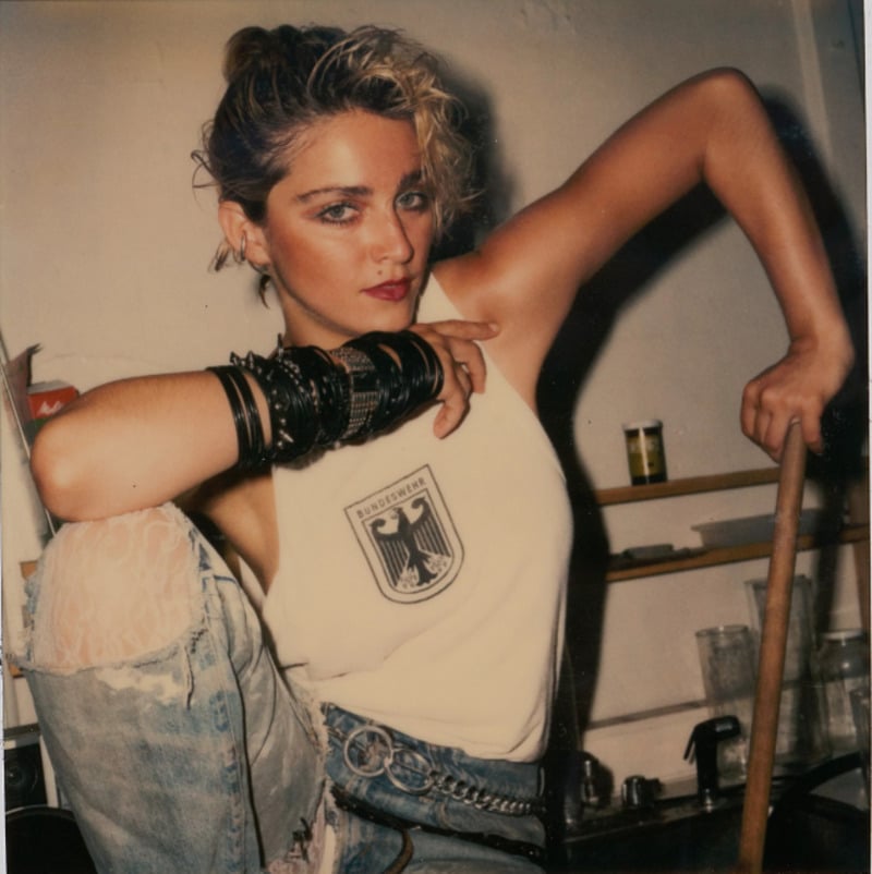 A woman with short blonde hair poses confidently, wearing a white tank top with a black eagle emblem, distressed jeans, and multiple black bracelets, sitting in a room with shelves and various objects in the background.
