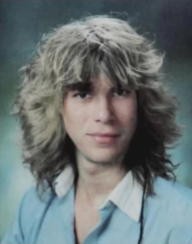 A young person with long, voluminous, wavy blond hair and a slight smile, wearing a light blue collared shirt, poses in front of a softly blurred background.
