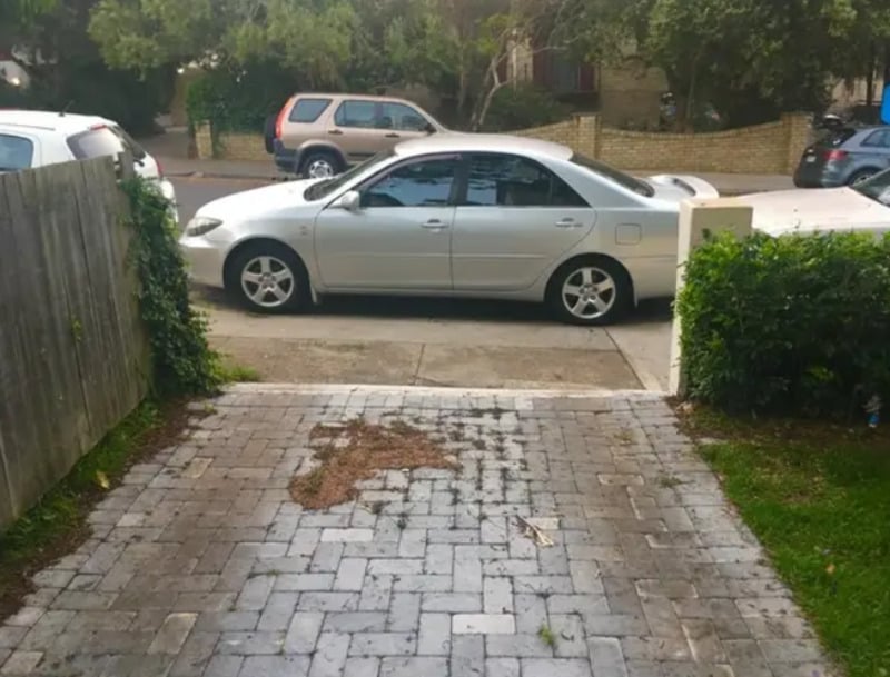 A silver sedan is parked across the end of a driveway, blocking the entrance. The driveway is paved with bricks and bordered by green grass and shrubs. Other cars and trees are visible in the background.