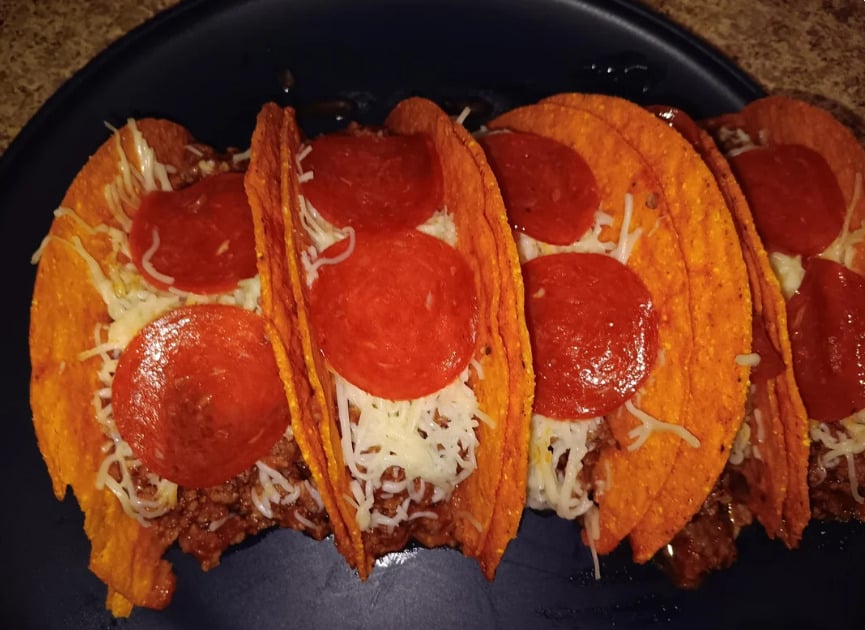 Four hard-shell tacos filled with ground meat, shredded cheese, and topped with pepperoni slices are arranged on a dark plate.