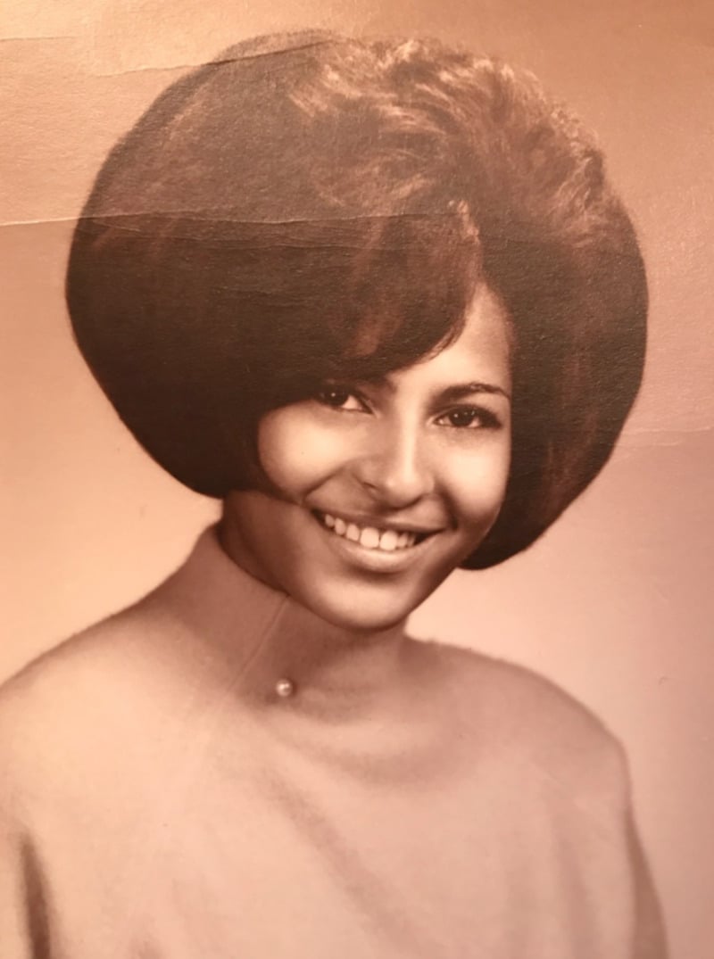 A young woman with a large, voluminous hairstyle smiles at the camera. She is wearing a light-colored, high-neck top with a small button detail. The photo is sepia-toned, giving it a vintage feel.