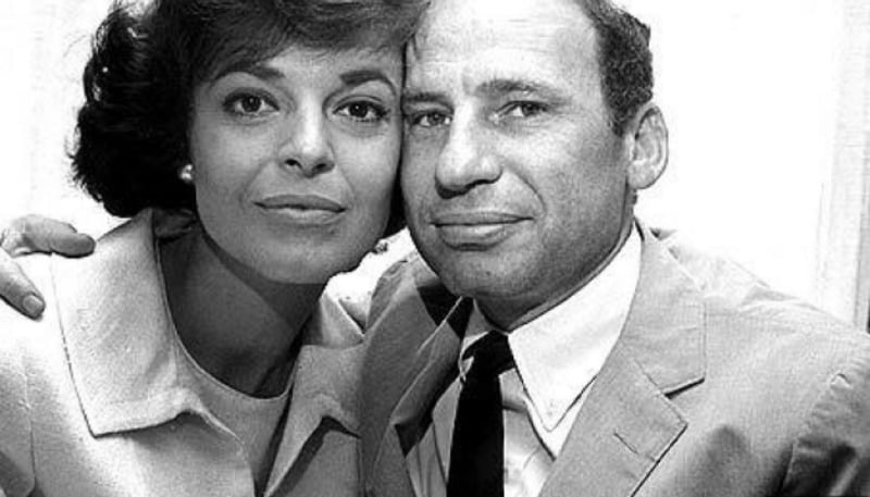 A black and white photo of a woman and a man posing closely together, both smiling slightly at the camera. The woman has short hair and wears a light-colored jacket. The man wears a suit and tie.