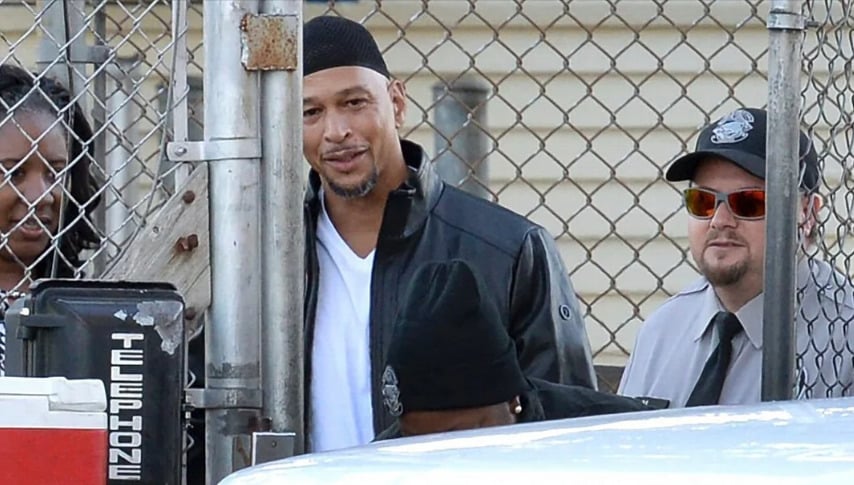A man in a black leather jacket and cap is smiling while standing by a chain-link fence; he is surrounded by others, including a security guard wearing sunglasses and a cap.