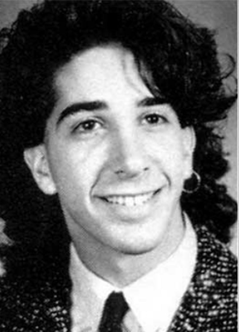 Black and white portrait of a young man with wavy dark hair, wearing a patterned blazer, white shirt, dark tie, and a hoop earring, smiling at the camera.