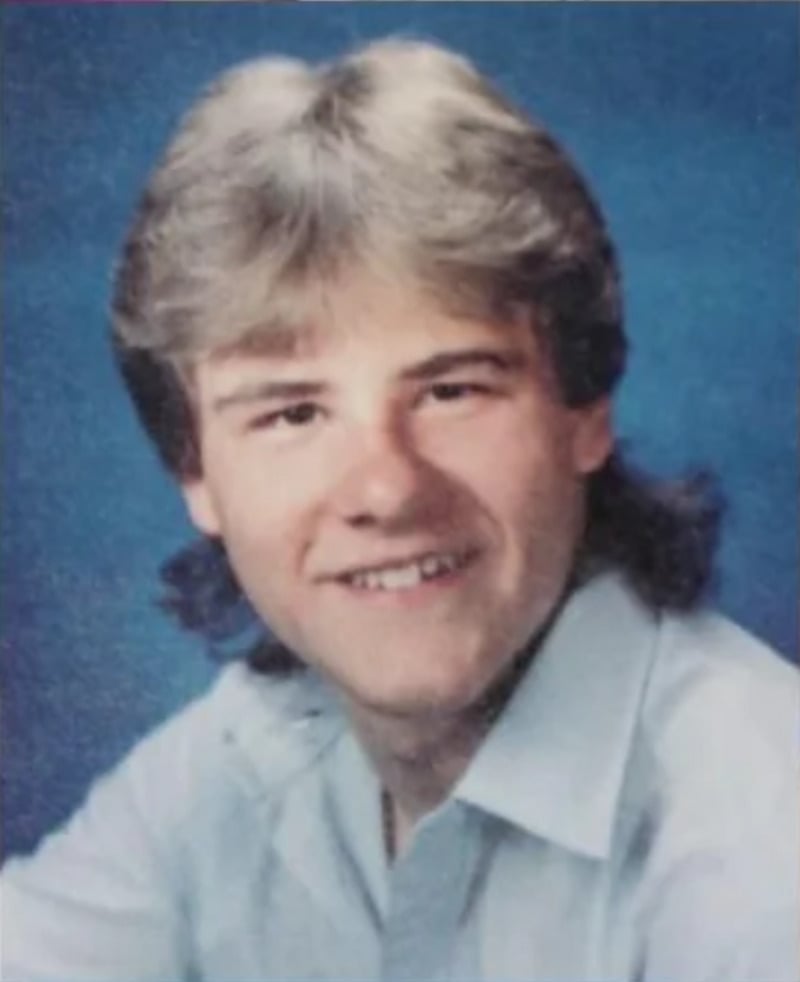 A young man with feathered blond hair and a mullet smiles at the camera, wearing a light blue collared shirt against a solid blue background, in a yearbook-style portrait.