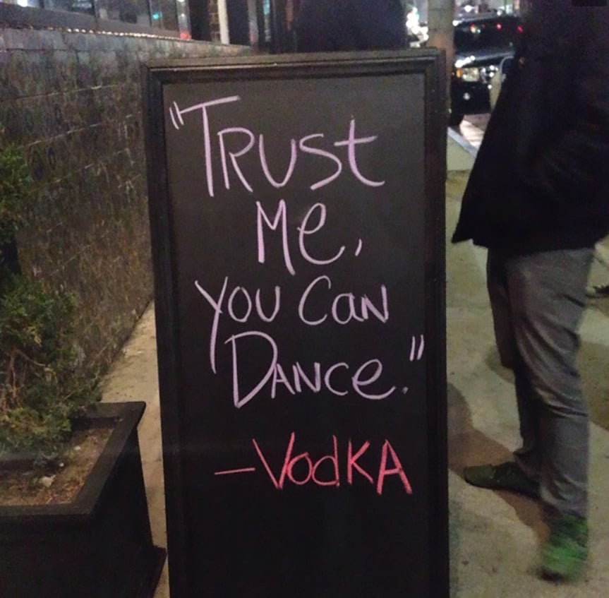 A chalkboard sign on a sidewalk reads, "'Trust me, you can dance.' - Vodka" in playful handwriting. A person stands nearby, and part of a plant and cars are visible in the background.