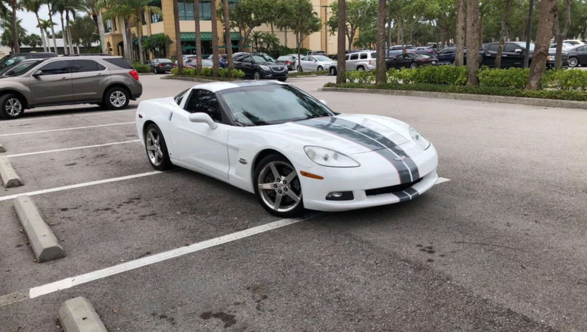 A white Chevrolet Corvette with black racing stripes is parked across two spaces in a parking lot surrounded by other cars and trees.