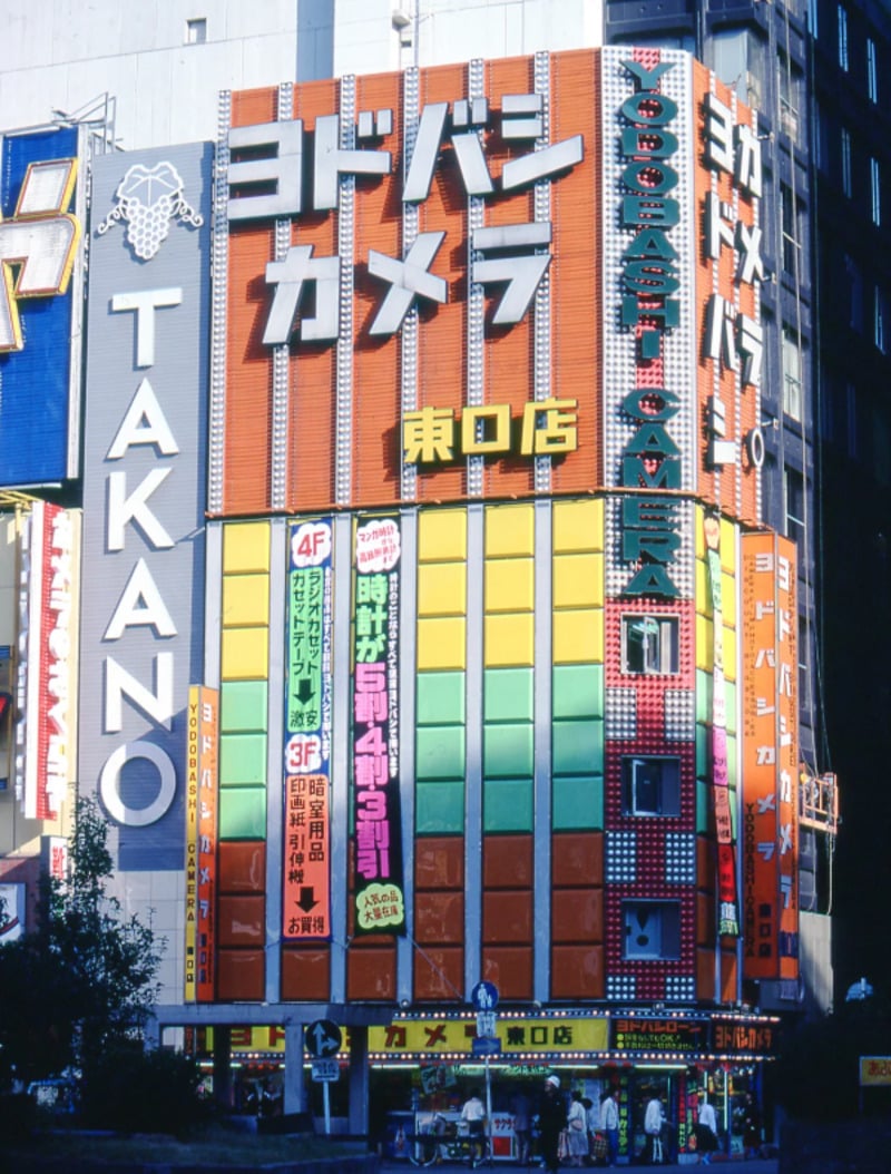 Colorful multi-story electronics store in Japan with large vertical signs in Japanese and English, including “Yodobashi Camera” and “TAKANO,” with vibrant banners and advertisements on the building facade.