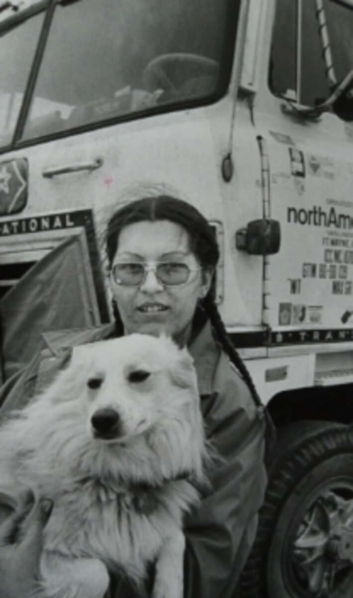 A person with glasses and braided hair holds a fluffy, light-colored dog in front of a large truck with "northAmerican" and other text visible on its side. The image is in black and white.