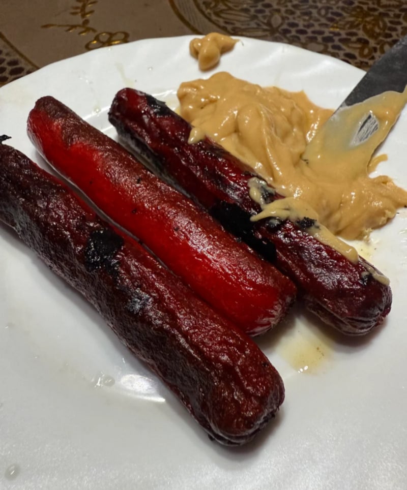 Three cooked red hotdogs on a white plate, with a generous serving of peanut butter and a knife beside them. The hotdogs appear slightly charred and shiny with oil.