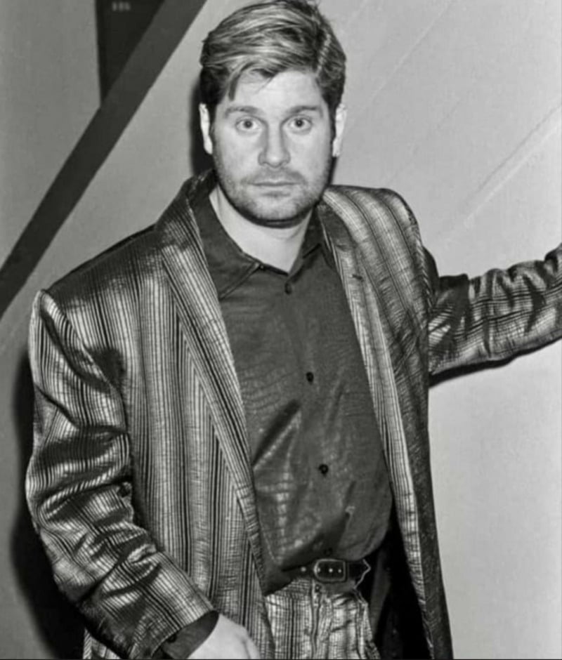 A man with short hair and a beard wears a shiny, striped suit jacket and matching pants, standing indoors with one arm resting on a wall, looking directly at the camera. The photo is in black and white.
