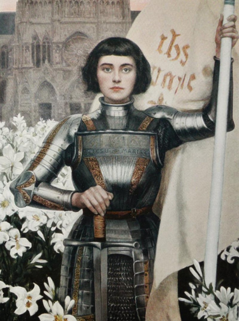 A woman in medieval armor stands among white lilies, holding a large white flag with gold writing. Behind her is a cathedral. Her expression is serious and determined.