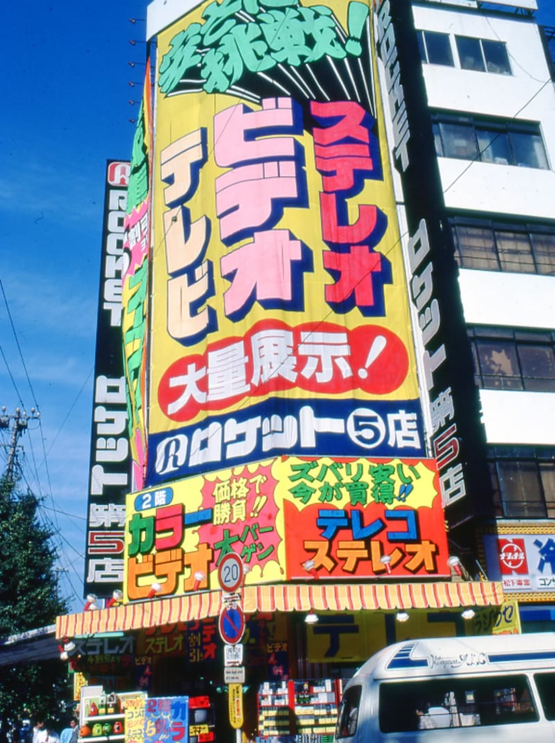 A tall building in Japan covered with large, colorful Japanese signs advertising electronics like stereo, video, and TV; street scene includes a van, pedestrians, and power lines under a blue sky.