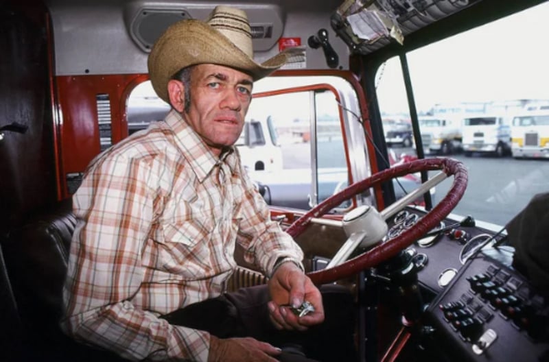 A man in a plaid shirt and cowboy hat sits in the driver’s seat of a large truck, holding small objects in his hand, with parking lot and vehicles visible outside the window.