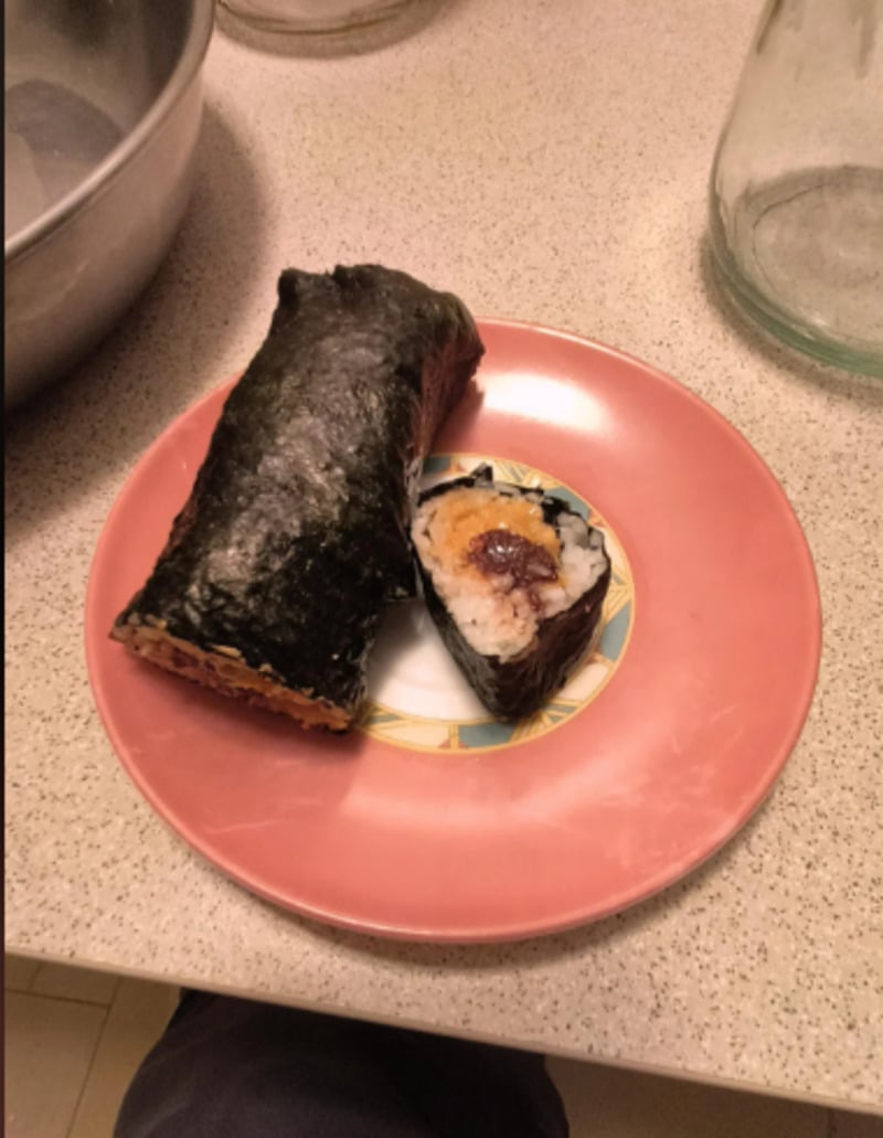 A large sushi roll wrapped in seaweed and a sliced piece showing rice and a dark filling are placed on a decorative plate with a pink rim, set on a kitchen counter.