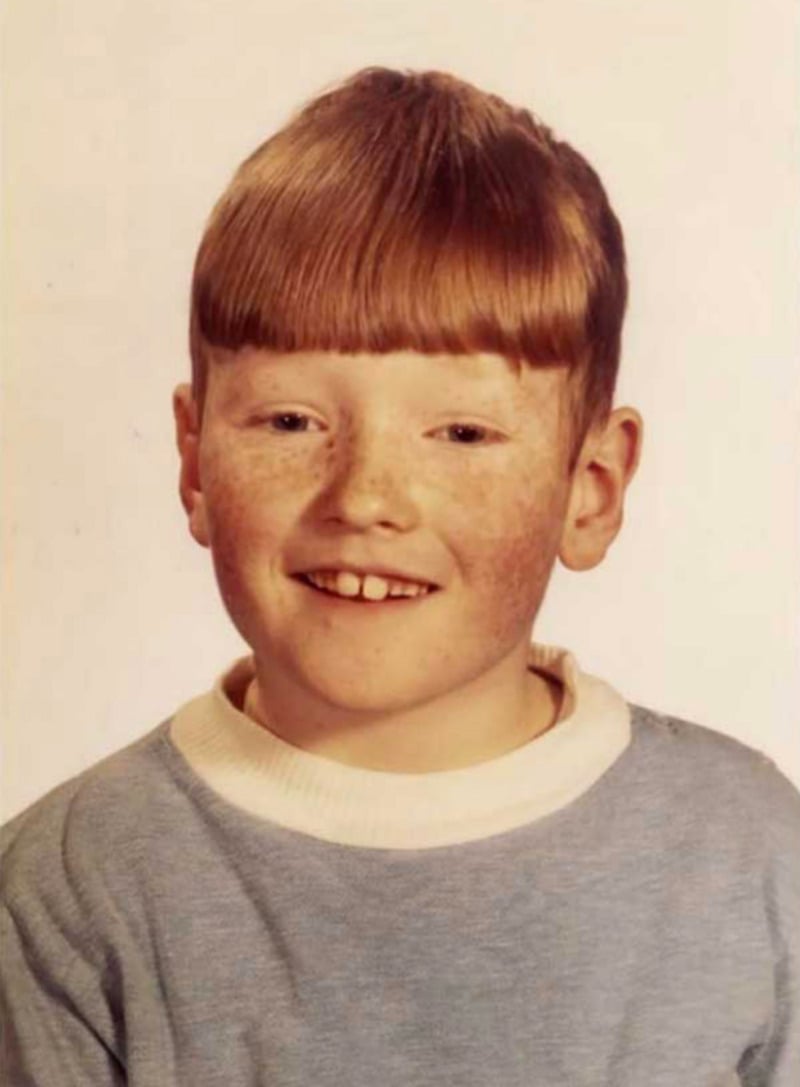 A young boy with red hair and freckles smiles at the camera. He is wearing a light blue sweater with a white collar. The background is plain and light-colored.
