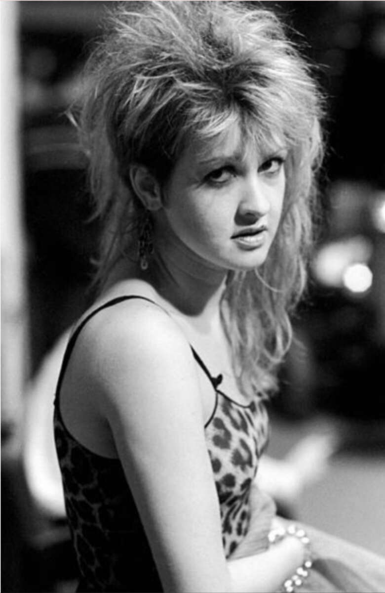 Black-and-white photo of a woman with voluminous, teased hair, wearing a leopard-print tank top and earrings, looking slightly over her shoulder with a serious expression.