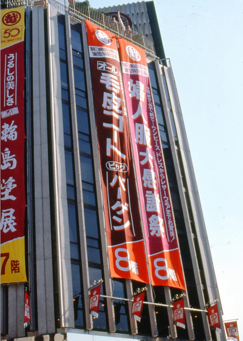 Tall building with large red vertical banners in Japanese advertising different events and products. Smaller flags with logos hang below the banners. The building has a modern design and metal railings at the top.