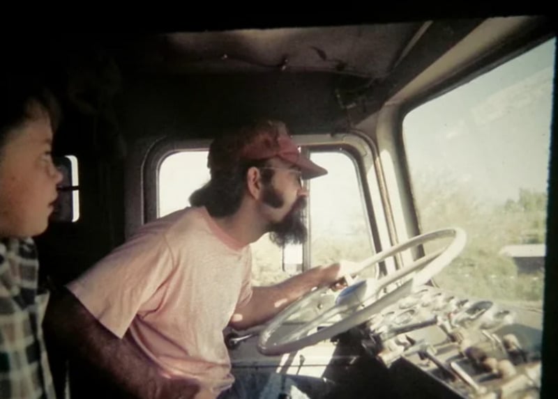 A man with a beard and glasses, wearing a red cap and pink shirt, leans forward while driving a large vehicle; a child sits beside him, looking ahead. Sunlight streams through the windows.