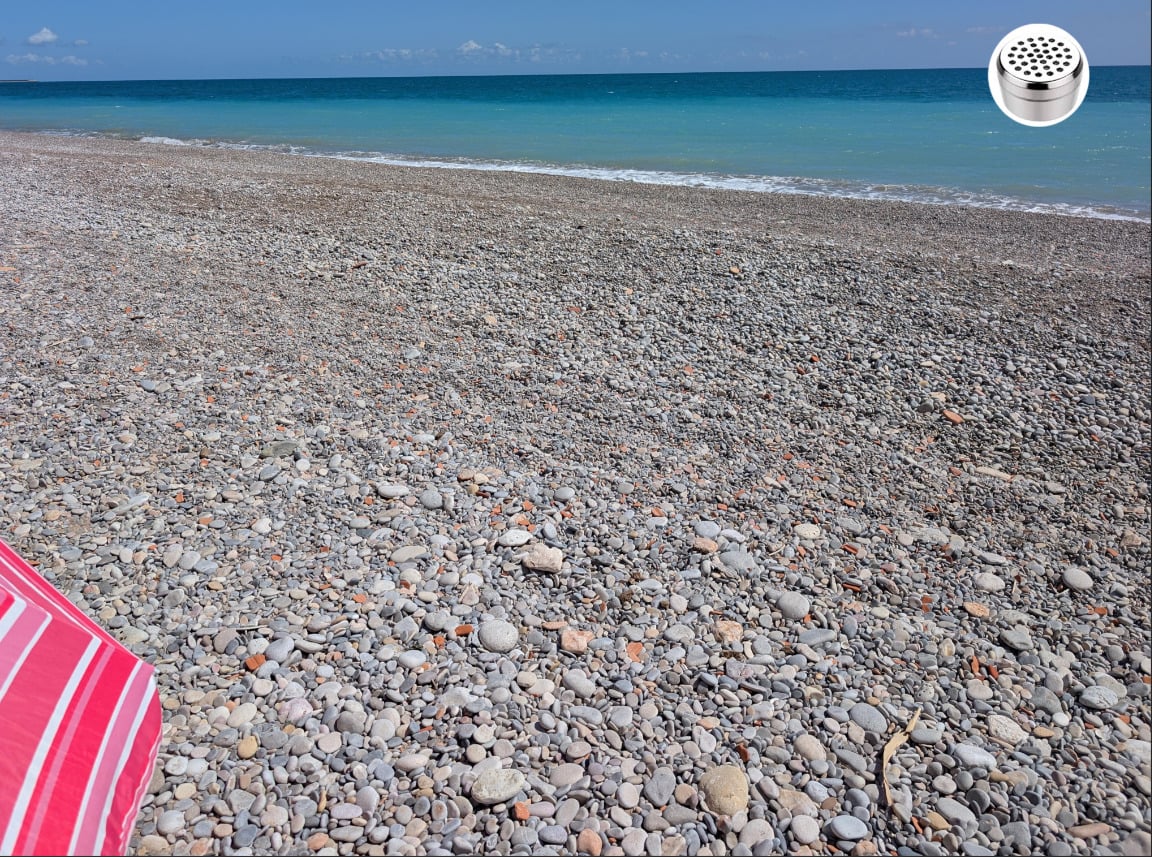A pebble-covered beach with blue-green sea and clear sky; a pink-striped umbrella is visible in the lower left corner. A small metal mesh object is in the upper right corner of the image.