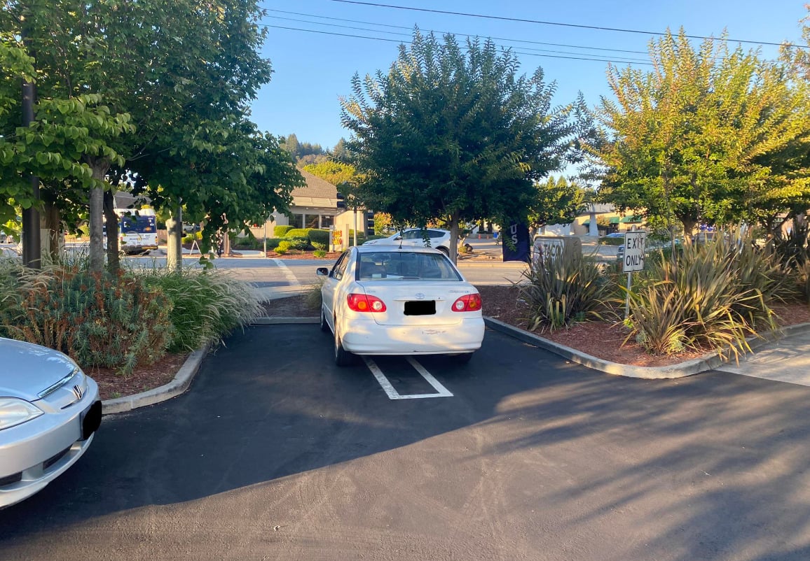 A white sedan is parked over the line in a parking lot exit lane, blocking part of the road. Trees and bushes line the sides, and a sign reads "EXIT ONLY." The sun is shining, casting shadows on the pavement.