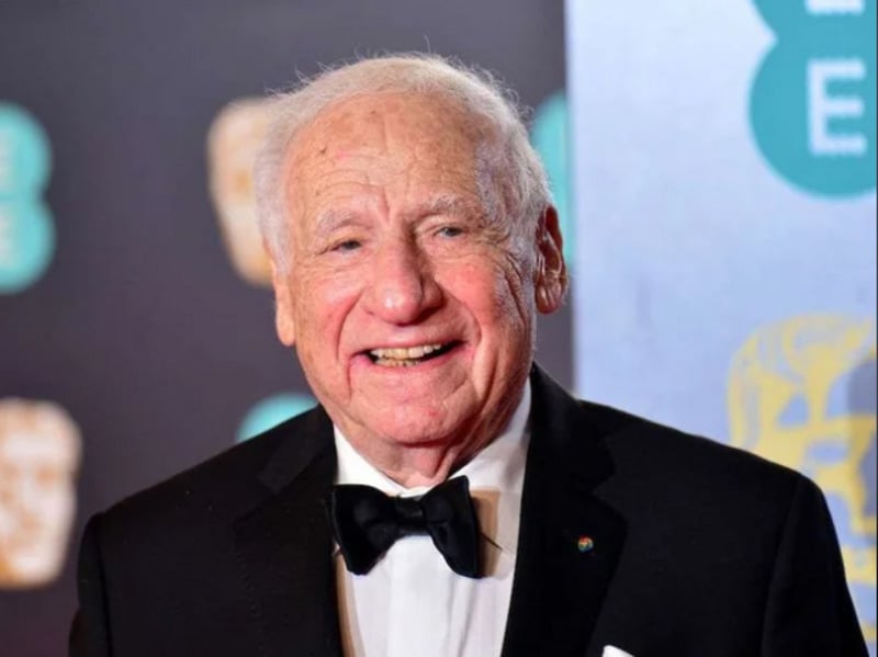 An elderly man in a tuxedo and bow tie smiles while standing in front of a blurred background with logos, likely at a formal event or award ceremony.